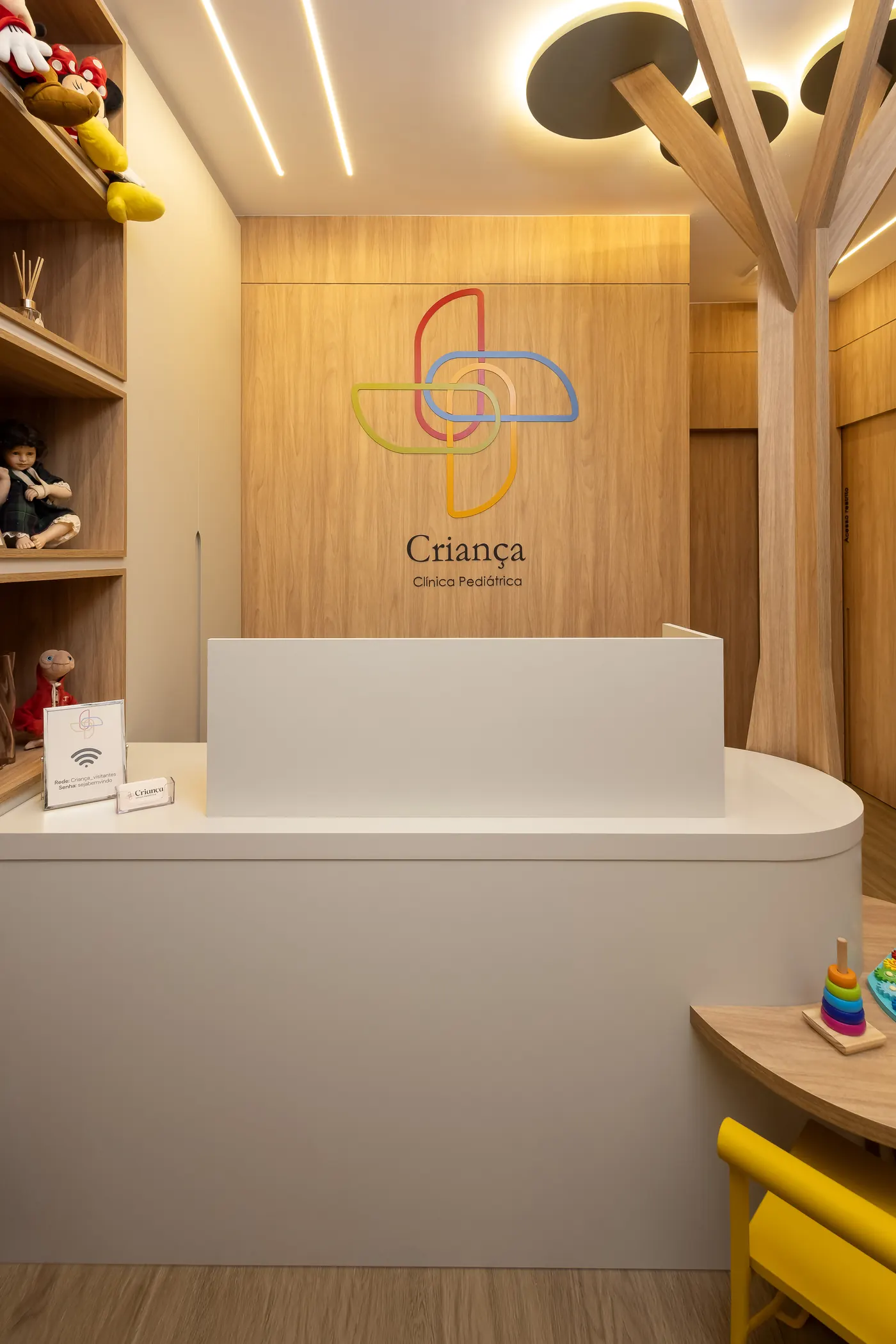 Recepção da Criança Clínica Pediátrica, com marcenaria em madeira clara e luminária em forma de árvore